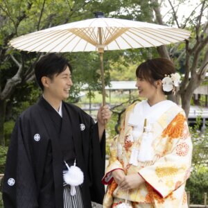 白い和傘をさして見つめ合う黒紋付袴の新郎と、白い髪飾りに金色の色打掛の花嫁。日本庭園の池のほとりで撮る和装前撮り