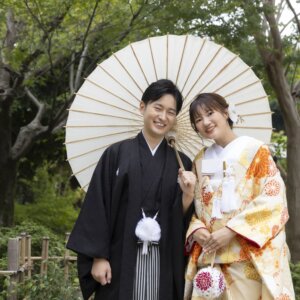 白い和傘の下で肩を寄せ、笑顔で並ぶ黒紋付袴の新郎と菊柄の色打掛の花嫁。白鳥庭園で撮る和装前撮りの和傘ショット