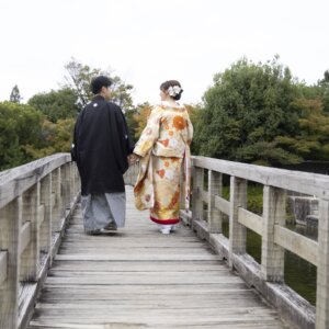 白鳥庭園の池にかかる木の橋を手をつないで渡る紋付袴の新郎と色打掛の花嫁の後ろ姿。日本庭園で撮る和装ロケーションフォト