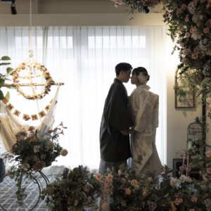フラワーウォールの前で白無垢の花嫁を抱き寄せ、顔を近づける羽織袴の新郎。洋風のスタジオセットで撮る和装前撮り