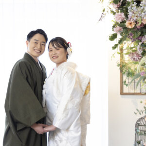 白いカーテン越しの光の中で、手を取り合って寄り添う羽織袴の新郎と白無垢の花嫁。フラワーウォールが映える名古屋のスタジオでの結婚写真