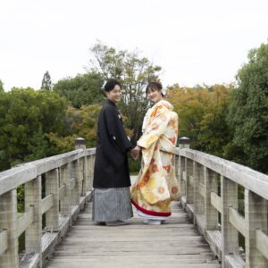 木の橋の上で振り返り、手をつないで微笑む縞袴の新郎と金色の色打掛の花嫁。白鳥庭園の橋で撮る和装前撮り