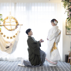 片膝をついてリングボックスを差し出す羽織袴の新郎と、驚く白無垢の花嫁。フラワーウォールの前で撮るプロポーズの和装前撮り