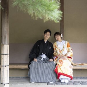 松の枝が垂れる茶室のベンチに並んで座り、笑顔を見せる黒紋付袴の新郎と色打掛の花嫁。白鳥庭園の和装前撮り