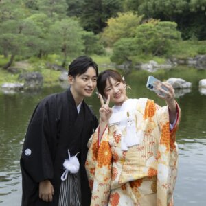 池を背にスマートフォンで自撮りをする、ピースサインの紋付袴の新郎と色打掛の花嫁。日本庭園で楽しむ和装前撮りの一コマ