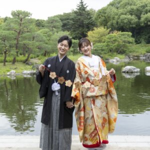 池のほとりで黄色い花のガーランドを手に並ぶ縞袴の新郎と菊柄の色打掛の花嫁。白鳥庭園で撮る秋の和装前撮り