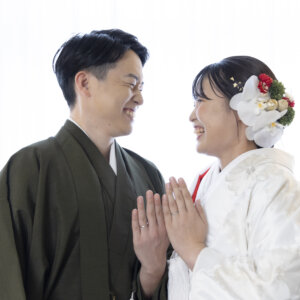 指輪をつけた手を重ね、顔を見合わせて笑う新郎と白無垢の花嫁。胡蝶蘭と赤い花の髪飾りが映える名古屋のスタジオでの和装前撮り