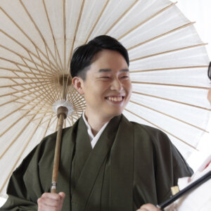 白い和傘の下で、花嫁に笑顔を向けるグリーンの羽織袴の新郎。和傘のアイテムで華やぐ名古屋のスタジオフォト