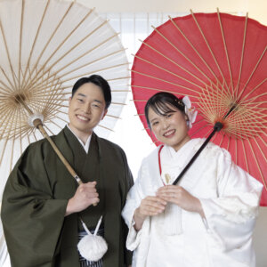 白と赤の和傘を肩にのせて並び、笑顔を見せる羽織袴の新郎と白無垢の花嫁。洋風のスタジオセットで撮る和装の結婚写真