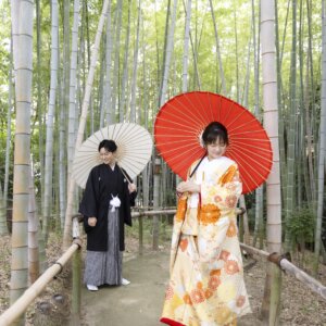 竹林の小道で白と赤の和傘をさして並ぶ黒紋付袴の新郎と金色の色打掛の花嫁。竹林で撮る和装前撮りの和傘ショット