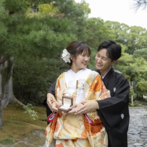 松の木の下でリングボックスを開き、後ろから花嫁を抱き寄せる黒紋付袴の新郎と色打掛の花嫁。白鳥庭園で撮るプロポーズ再現の和装前撮り