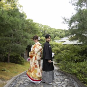 松と苔の庭園の石畳の小道で振り返る、金色の色打掛の花嫁と黒紋付袴の新郎。日本庭園で撮る色打掛の和装ロケーションフォト