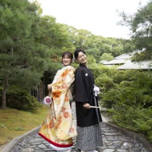 庭園の石畳の小道で手をつなぎ、振り返って微笑む縞袴の新郎と菊柄の色打掛の花嫁。白鳥庭園で撮る秋の和装前撮り