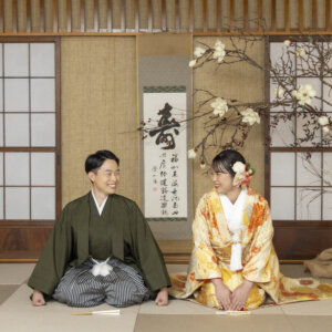 障子と寿の掛け軸の前で正座し、笑顔で顔を見合わせる羽織袴の新郎と山吹色の色打掛の花嫁。和室で撮る和装前撮り