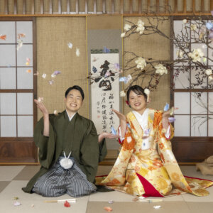 寿の掛け軸と白い花木を飾った和室のスタジオで、折り鶴を舞わせて笑うグリーンの羽織袴の新郎と色打掛の花嫁。名古屋のスタジオで撮る和装前撮り