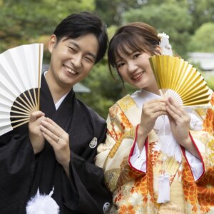 白い扇子と金色の扇子で顔を半分隠して笑う黒紋付袴の新郎と色打掛の花嫁。扇子を使った和装前撮りの定番ポーズ