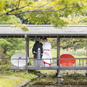 池にかかる白鳥庭園の屋根付きの渡り廊下で、白と赤の和傘を手に並ぶ黒紋付袴の新郎と白無垢の花嫁。日本庭園で撮る和装前撮り