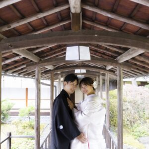 木の梁が連なる渡り廊下で、頬を寄せ合って微笑む紋付袴の新郎と白無垢の花嫁。白鳥庭園の和装前撮りで人気のポーズ
