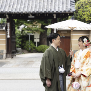 瓦屋根の門の前で、白い和傘を差して笑い合う羽織袴の新郎と色打掛の花嫁。名古屋の街なかで撮る和装のロケーションフォト