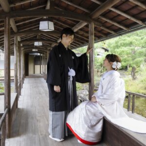 渡り廊下の縁に座った白無垢の花嫁の綿帽子にそっと手を添える黒紋付袴の新郎。白鳥庭園の茶室で撮る和装前撮り