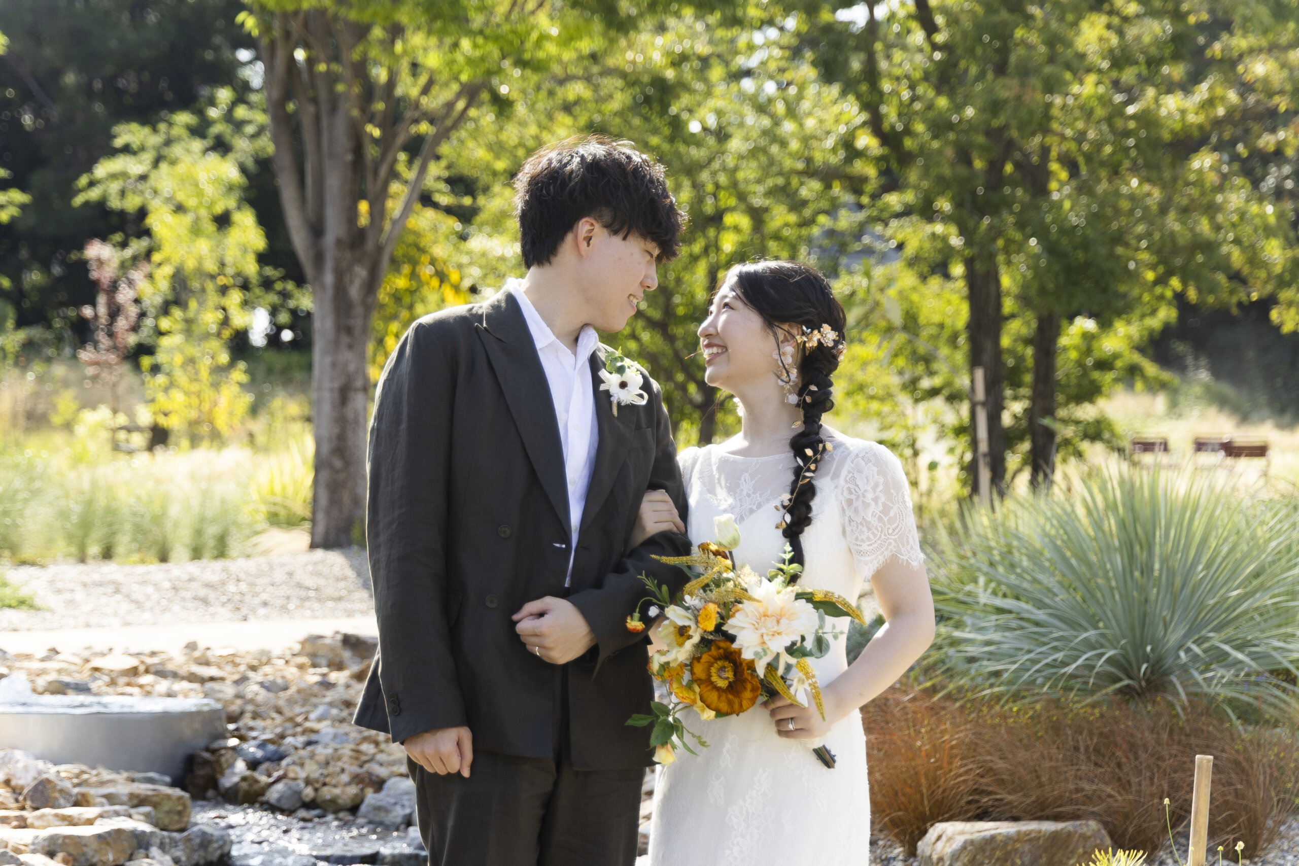 木漏れ日の庭園で腕を組んで見つめ合う新郎新婦と、イエローのブーケ