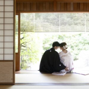 障子を開け放った茶室の縁側に並んで座り、庭の緑を眺める紋付袴の新郎と白無垢の花嫁の後ろ姿。和室で撮る和装前撮り