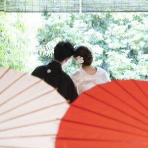 白と赤の和傘を手前に置き、縁側で庭を眺める新郎と白無垢の花嫁の後ろ姿。白鳥庭園の茶室で撮る和傘を使った和装前撮り