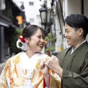 路地の店先で、小指を絡めて笑い合う色打掛の花嫁とグリーンの羽織袴の新郎。指切りのポーズが楽しい和装の結婚写真