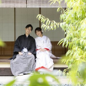 新緑の葉越しに見える、茶室の縁側に座って微笑む縞袴の新郎と白無垢の花嫁。白鳥庭園の日本庭園で撮る和装前撮り