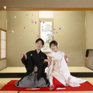 掛け軸のある和室で、折り鶴を空に舞わせて笑う縞袴の新郎と白無垢の花嫁。白鳥庭園の茶室で楽しむ折り鶴の和装前撮り