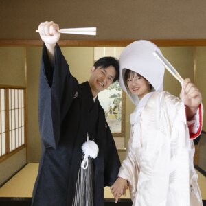 和室で末広を高く掲げて笑う紋付袴の新郎と、綿帽子の白無垢の花嫁。白鳥庭園の茶室で撮る和装前撮りの楽しい一枚
