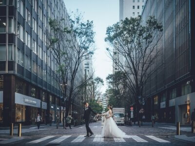 並木が続く東京のオフィス街の横断歩道で、手をつないで歩くタキシードの新郎とウェディングドレスの花嫁のロケーションフォト