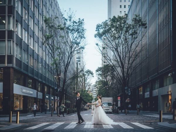並木が続く東京のオフィス街の横断歩道で、手をつないで歩くタキシードの新郎とウェディングドレスの花嫁のロケーションフォト