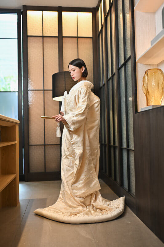 格子窓から光が差し込む和モダンな部屋で、扇子を手に横を向いて裾を引く白無垢の花嫁。白無垢AB102の後ろ姿の柄も見える和装前撮り