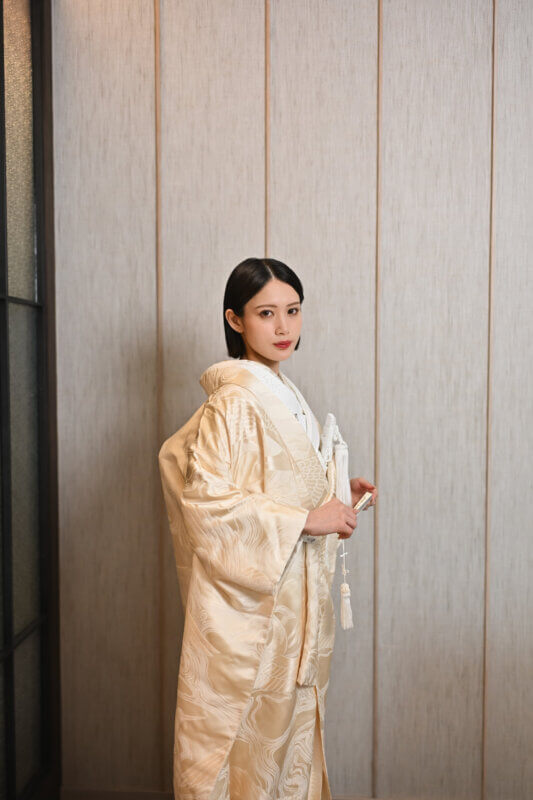 木目の壁の前で扇子と房飾りを持ち、正面を向いて立つ金糸の刺繍入りの白無垢の花嫁。スタジオフォトで撮影した衣装写真